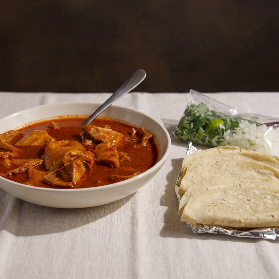 Menudo in a white bowl with a spoon, alongside tortillas, chopped onions, and cilantro on a white tablecloth.